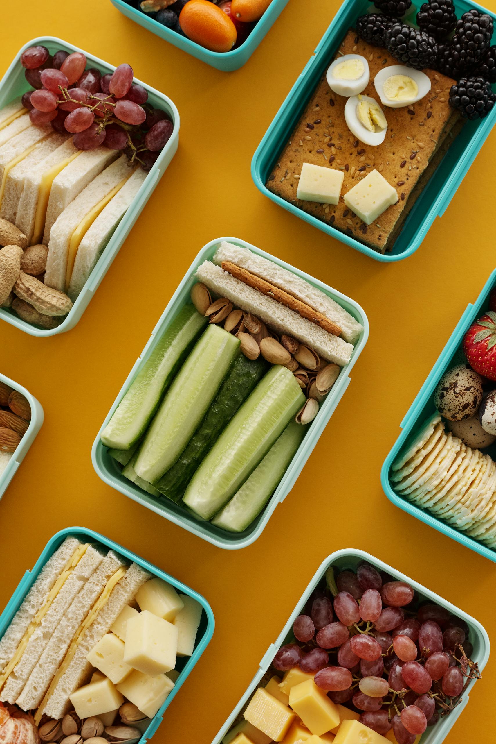 Photo by Vanessa Loring | Motherhues An assortment of colorful lunch boxes with fruits, vegetables, and snacks on a vibrant yellow background.