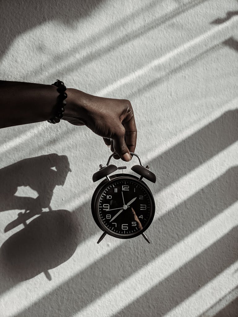 Photo by Ola Dapo | Motherhues Close-up of a hand holding a vintage alarm clock casting shadows in daylight.