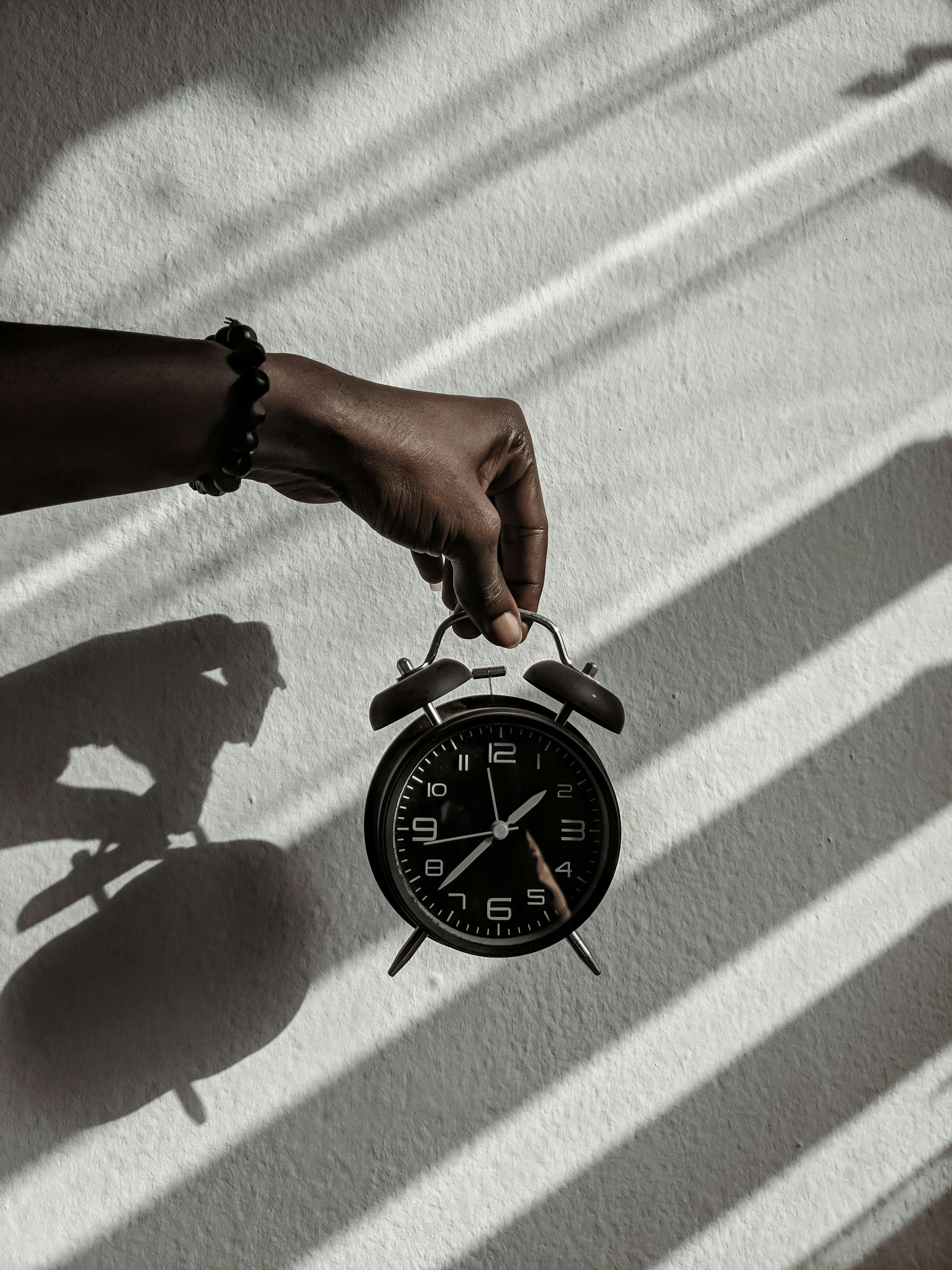 Photo by Ola Dapo | Motherhues Close-up of a hand holding a vintage alarm clock casting shadows in daylight.