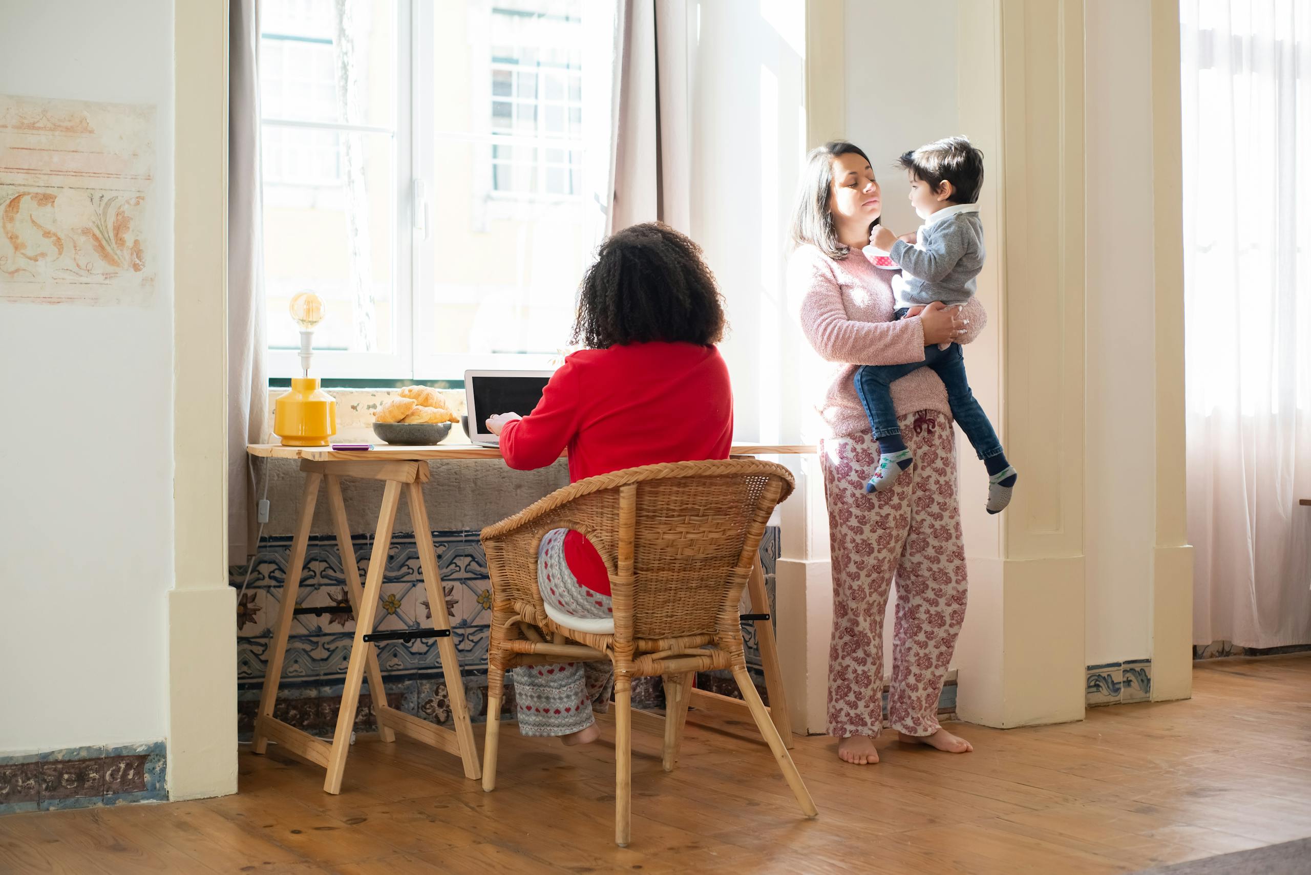 A mom holds her child while another person works on a laptop in a sunlit, cozy living room.