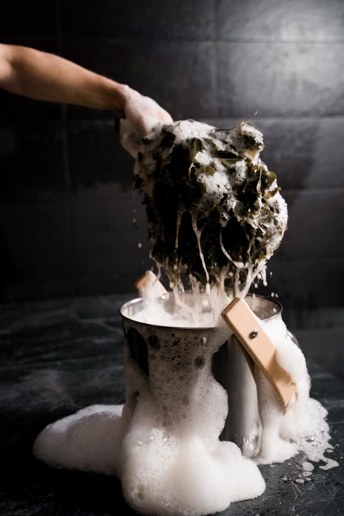 Photo by Ron Lach | Motherhues A hand scrubs baseball uniform over a bucket with soap suds and foam, highlighting cleaning and hygiene.