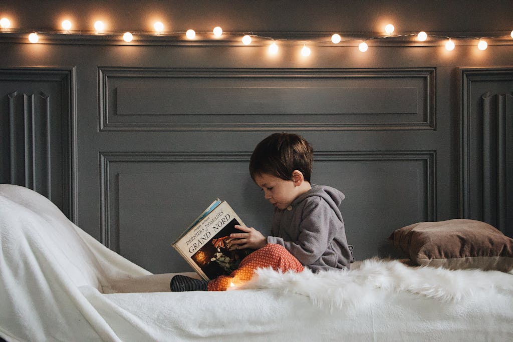 Photo by Maël BALLAND | Motherhues A young boy reads a book in a cozy bedroom setting with warm string lights and soft textures.