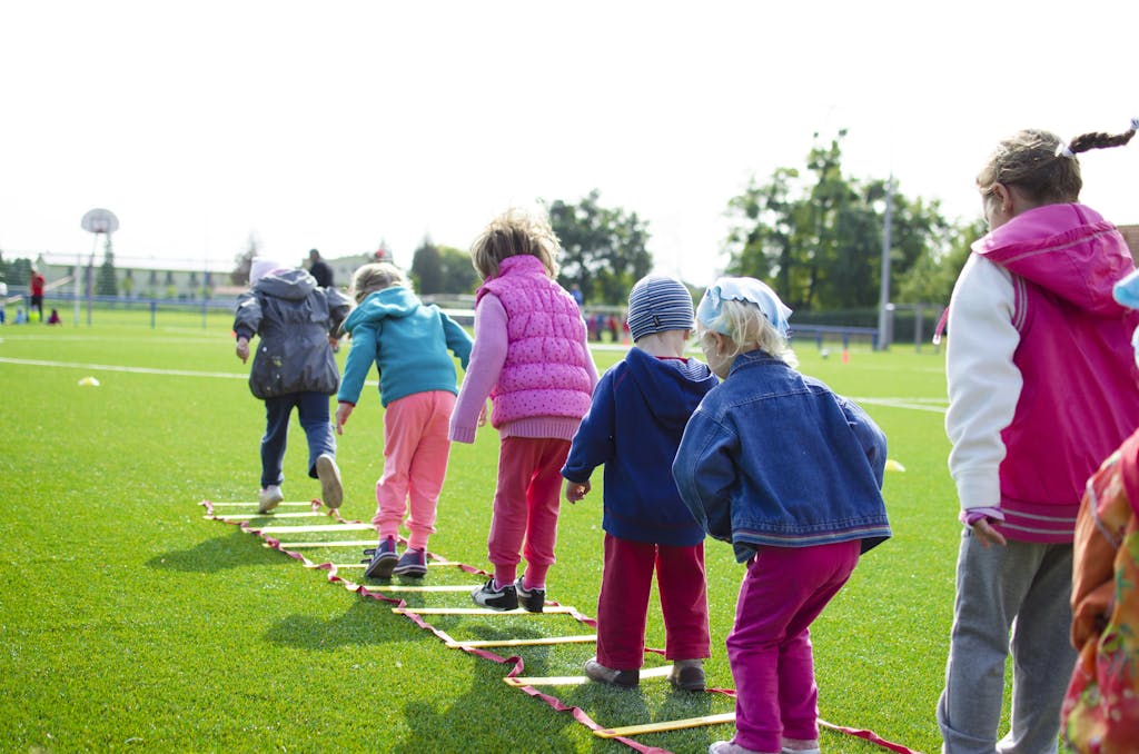 Photo by Lukas | Motherhues Kids enjoy an outdoor activity on a grassy field, stepping over a ladder.