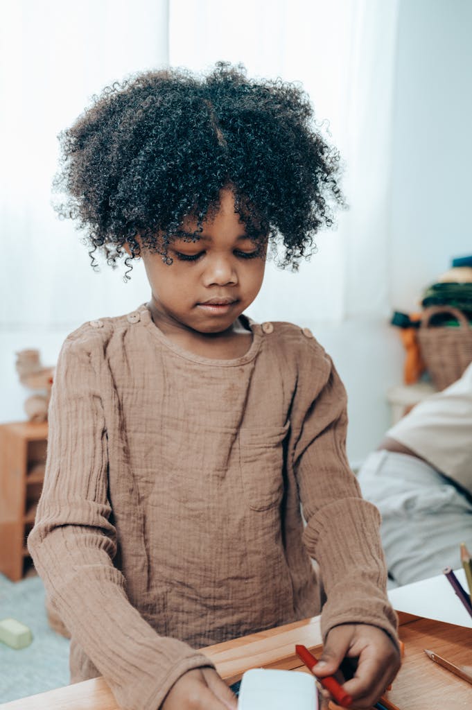 Photo by Ketut Subiyanto | Motherhues From above of adorable pensive black kid with Afro hairstyle in casual clothes sitting with white paper cube and red felt pen at wooden table at home and looking down