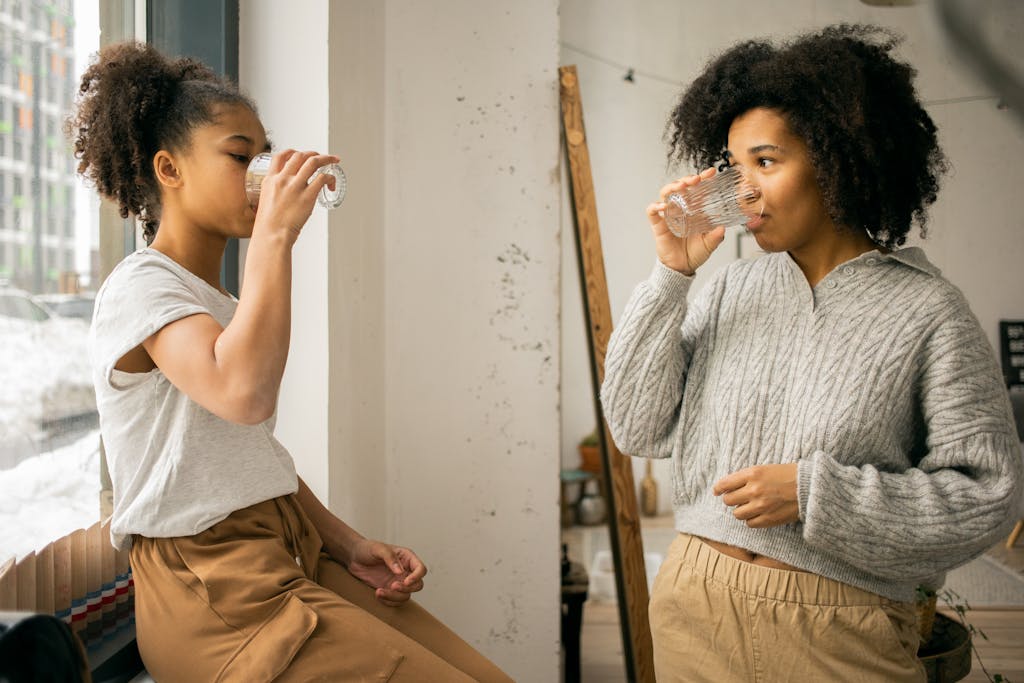 Photo by Monstera Production | Motherhues Side view of African American daughter and mother in casual wear drinking water from glasses near window in light room