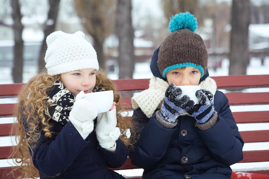 Photo by Aleks | Motherhues Two children bundled in winter clothing enjoy a warm drink on a snowy bench.