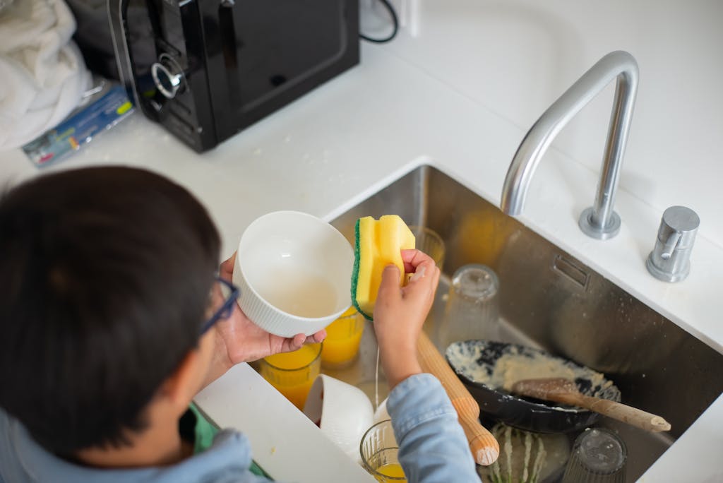 Photo by Kampus Production | Motherhues A young boy diligently washing dishes with a sponge in the kitchen sink.