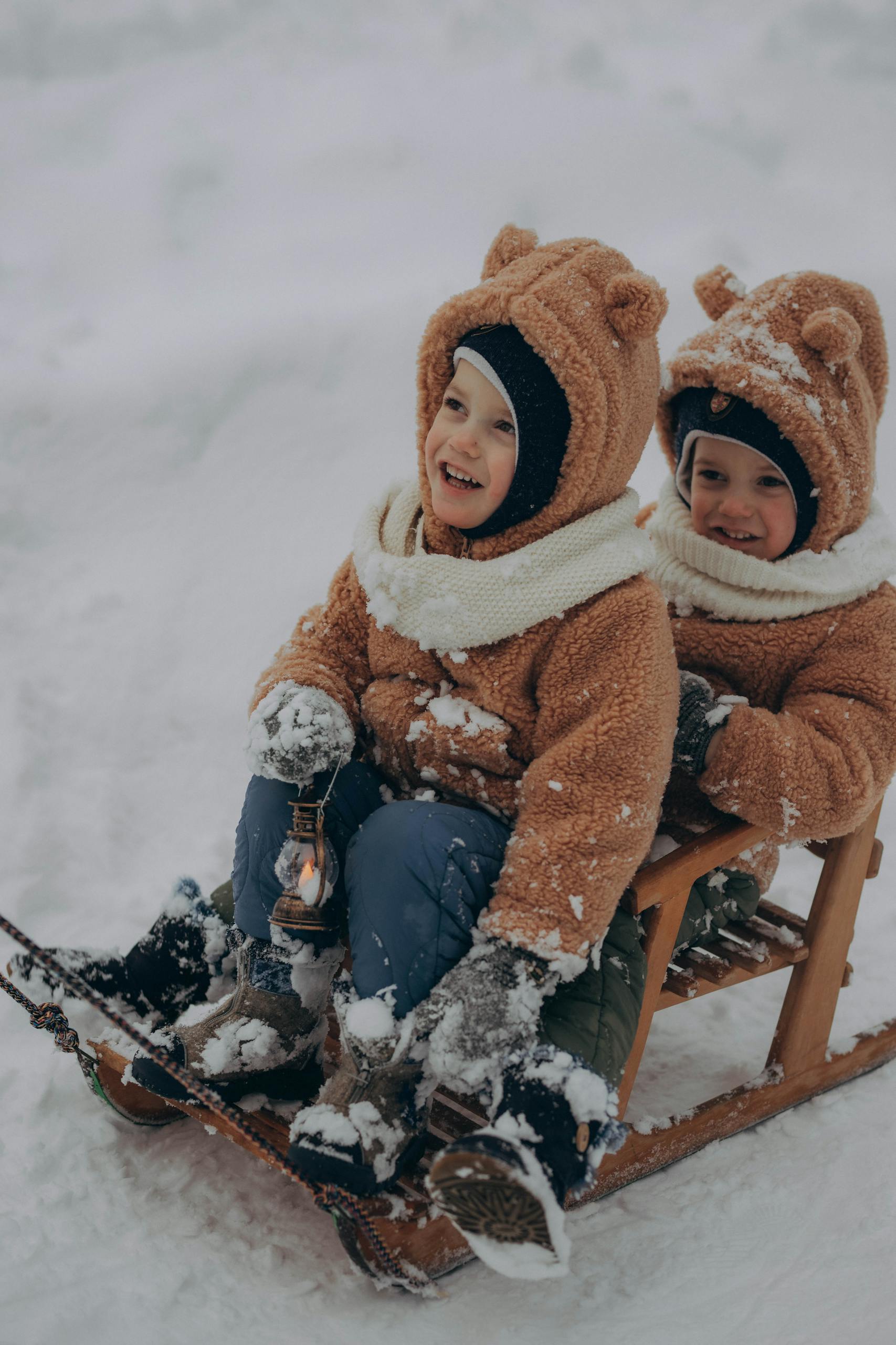 Two smiling children in warm clothing having fun in the snow on a wooden sled.