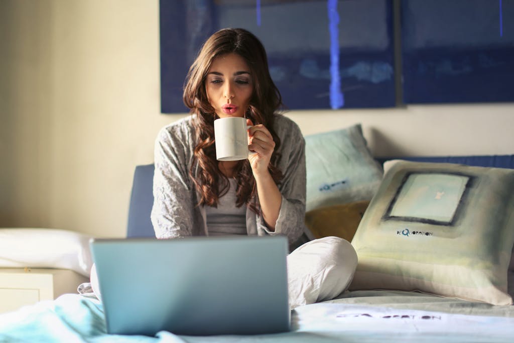 Photo by Andrea Piacquadio | Motherhues A woman enjoying coffee while working from home routine in a cozy bedroom setting with a laptop.